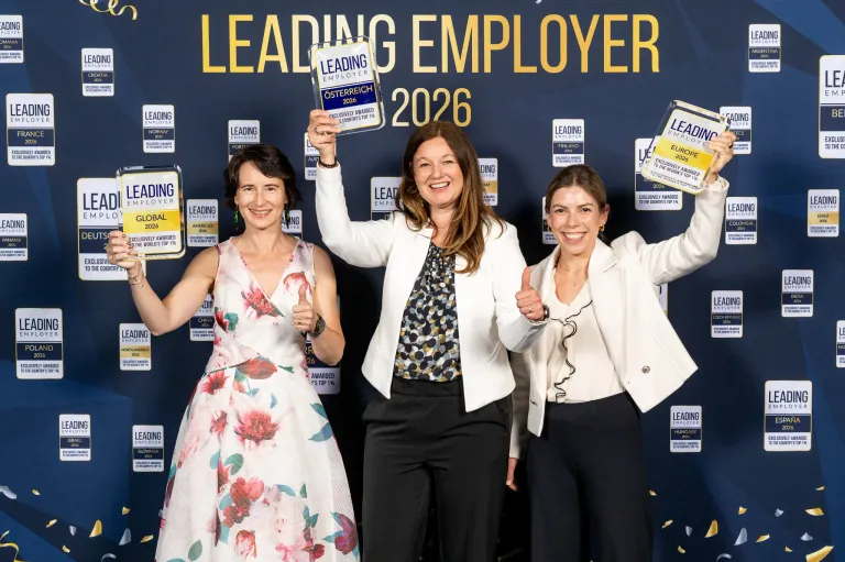Drei Frauen stehen zusammen und halten Auszeichnungen. Eine hält ein Schild mit der Aufschrift 'Führender Arbeitgeber 2026'. Die Frau in der Mitte trägt eine weiße Jacke und schwarze Hose. Die Frau auf der linken Seite trägt ein Blumenkleid. Die Frau auf der rechten Seite trägt eine weiße Jacke.