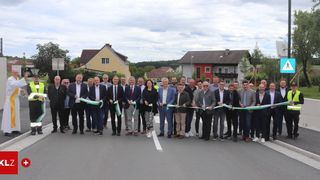 Eine Gruppe formell gekleideter Männer und Frauen steht mit grünen Bändern auf einer Straße, wahrscheinlich zur Feier eines Bandabschneidens.