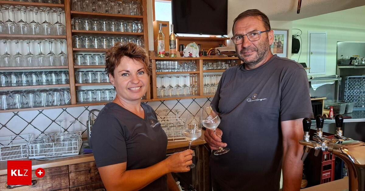 Ein lächelnder Mann und eine Frau stehen in einer Bar, halten Weingläser in den Händen, mit Regalen voller Gläser und Flaschen im Hintergrund.