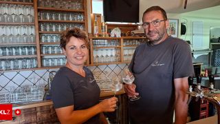 Ein lächelnder Mann und eine Frau stehen in einer Bar, halten Weingläser in den Händen, mit Regalen voller Gläser und Flaschen im Hintergrund.