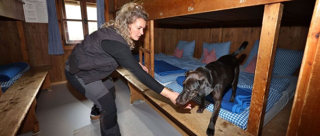 A woman pets her black dog on a bunk bed in a wooden room. She wears a vest and boots. Two beds with pillows are on either side.