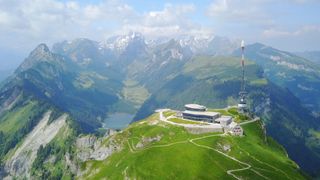 Luftaufnahme eines Gebäudes auf einem Berg mit einem Turm, umgeben von üppigem Grün und Bergen in der Ferne.