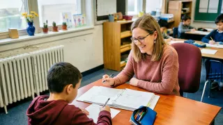 Eine lächelnde Frau mit Brille sitzt an einem Schreibtisch mit einem Jungen. Sie hält einen Stift und er schreibt in ein Notizbuch. Dahinter steht ein Holzregal mit verschiedenen Gegenständen.