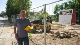 Ein Mann in einem grauen T-Shirt und Jeans steht vor einem Metallzaun. Dahinter steht ein Baufahrzeug, und ein Schild mit der Aufschrift 'REITER ERDBAU-KIES' ist zu sehen.