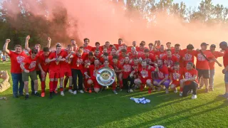 Eine Fußballmannschaft feiert mit einer Trophäe auf einem Feld, umgeben von rotem Rauch. Spieler in roten Uniformen und Medaillen posieren mit einem großen Silberpokal.