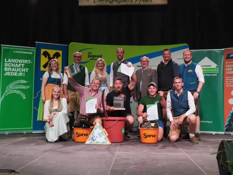 A group of people posing on stage, some holding certificates and buckets, smiling. They are dressed in traditional clothing. In the background, there is a green backdrop with logos and text.