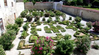 Ein formeller Garten mit komplexen Mustern aus grünen und rosa Pflanzen. Der Garten ist von Steinmauern umgeben und enthält ein weißes Pavillon.