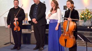 Vier Musiker, zwei mit Geigen und zwei mit Celli, stehen drinnen, möglicherweise in einer Kirche. Sie sind formell gekleidet.