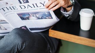Ein Mann liest eine Zeitung mit dem Titel 'The Daily' mit einem Bild einer Stadtsilhouette auf der Titelseite. Er trägt eine Uhr und sitzt in einem Stuhl in einem Café.