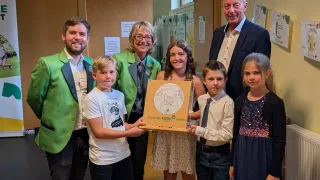 Eine Gruppe von Erwachsenen und Kindern steht zusammen und lächelt für ein Foto. Sie halten eine Box mit einer Zeichnung eines Hundes. Die Kinder tragen Freizeitkleidung. Die Frau in der grünen Jacke hält ein Papier mit einem Hund darauf. Hinter ihnen befinden sich Holzpaneele mit Postern.