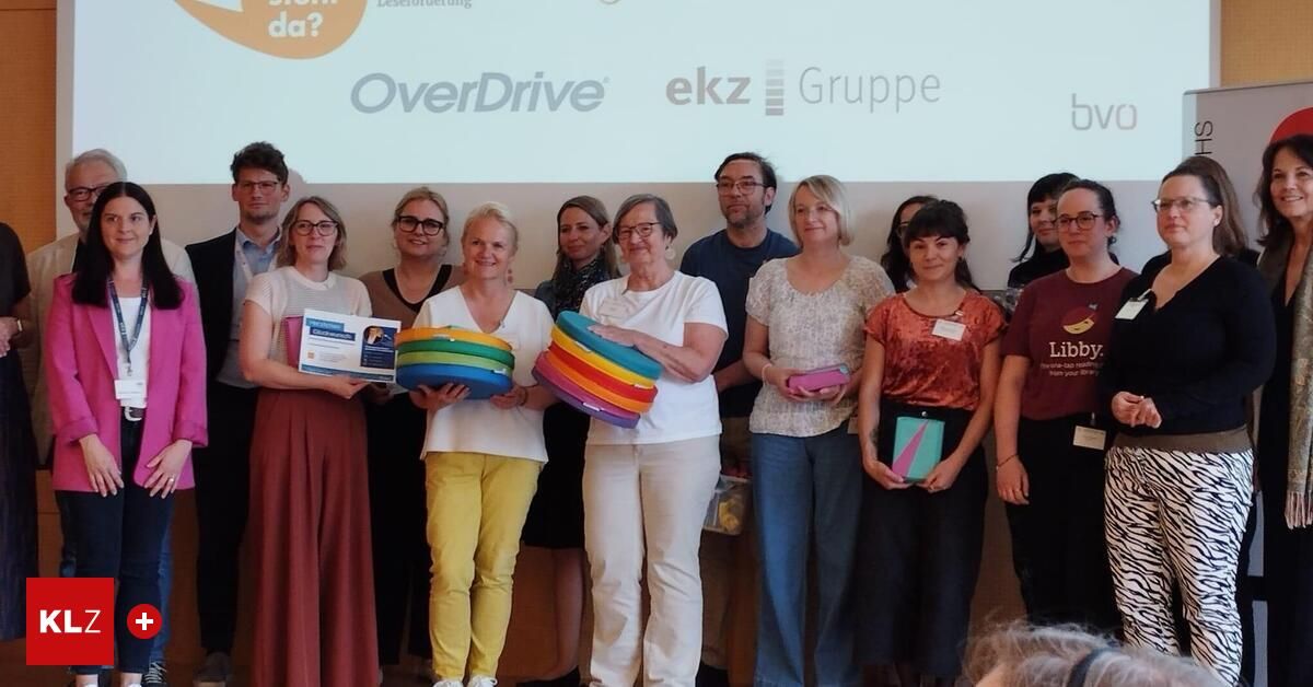 Eine Gruppe von Menschen posiert für ein Foto, hält bunte Gegenstände, wahrscheinlich Bücher, vor einer Projektionsleinwand mit den Logos 'OverDrive' und 'ekz Gruppe'.