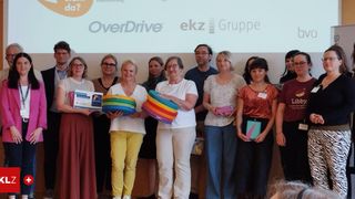 Eine Gruppe von Menschen posiert für ein Foto, hält bunte Gegenstände, wahrscheinlich Bücher, vor einer Projektionsleinwand mit den Logos 'OverDrive' und 'ekz Gruppe'.