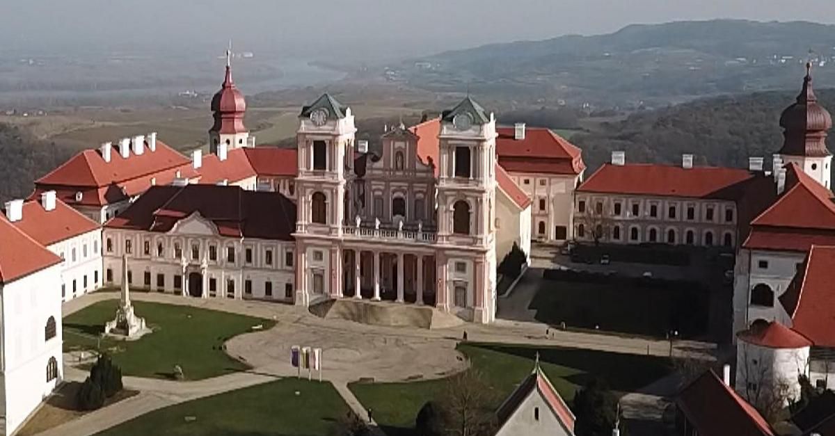 Ein großes Barockgebäude mit rotem Dach und weißen Wänden, umgeben von Gras und Bergen.