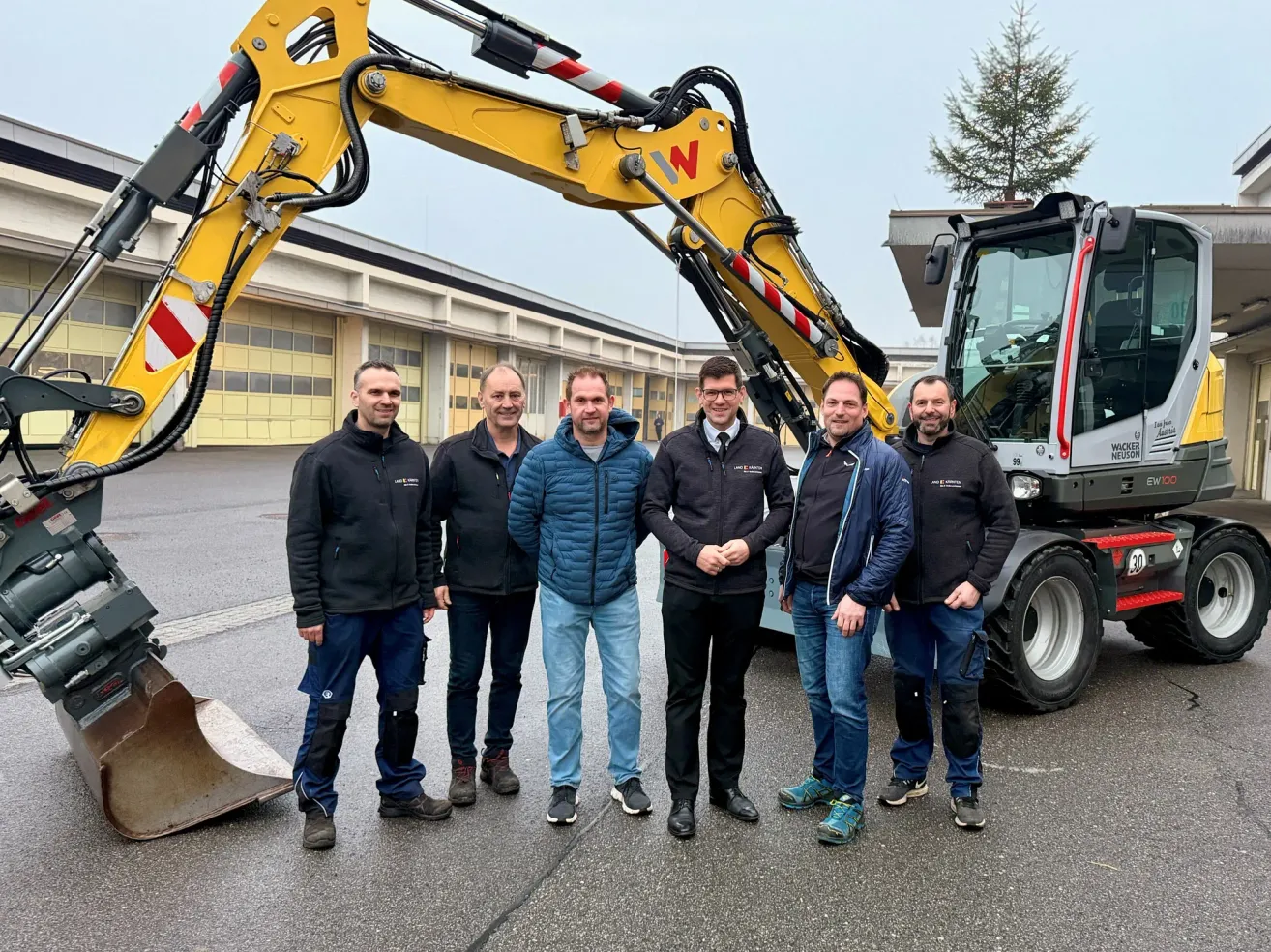Fünf Männer stehen vor einem großen gelben Baufahrzeug, wahrscheinlich einem Bagger, mit einem Gebäude im Hintergrund. Sie scheinen für ein Foto zu posieren.