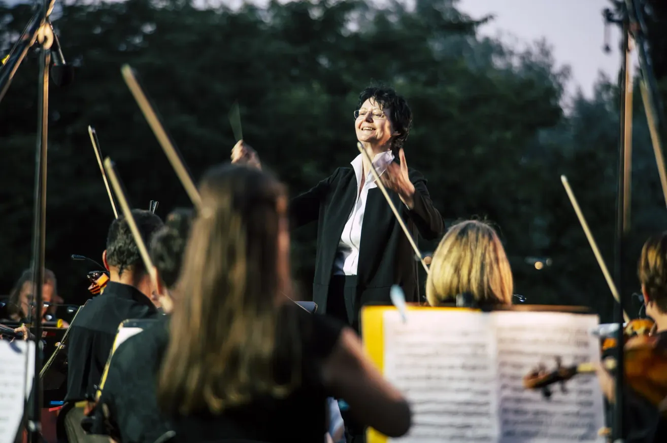 Eine Frau mit Brille dirigiert ein Orchester im Freien, umgeben von Musikern mit Musikinstrumenten und Notenblättern.