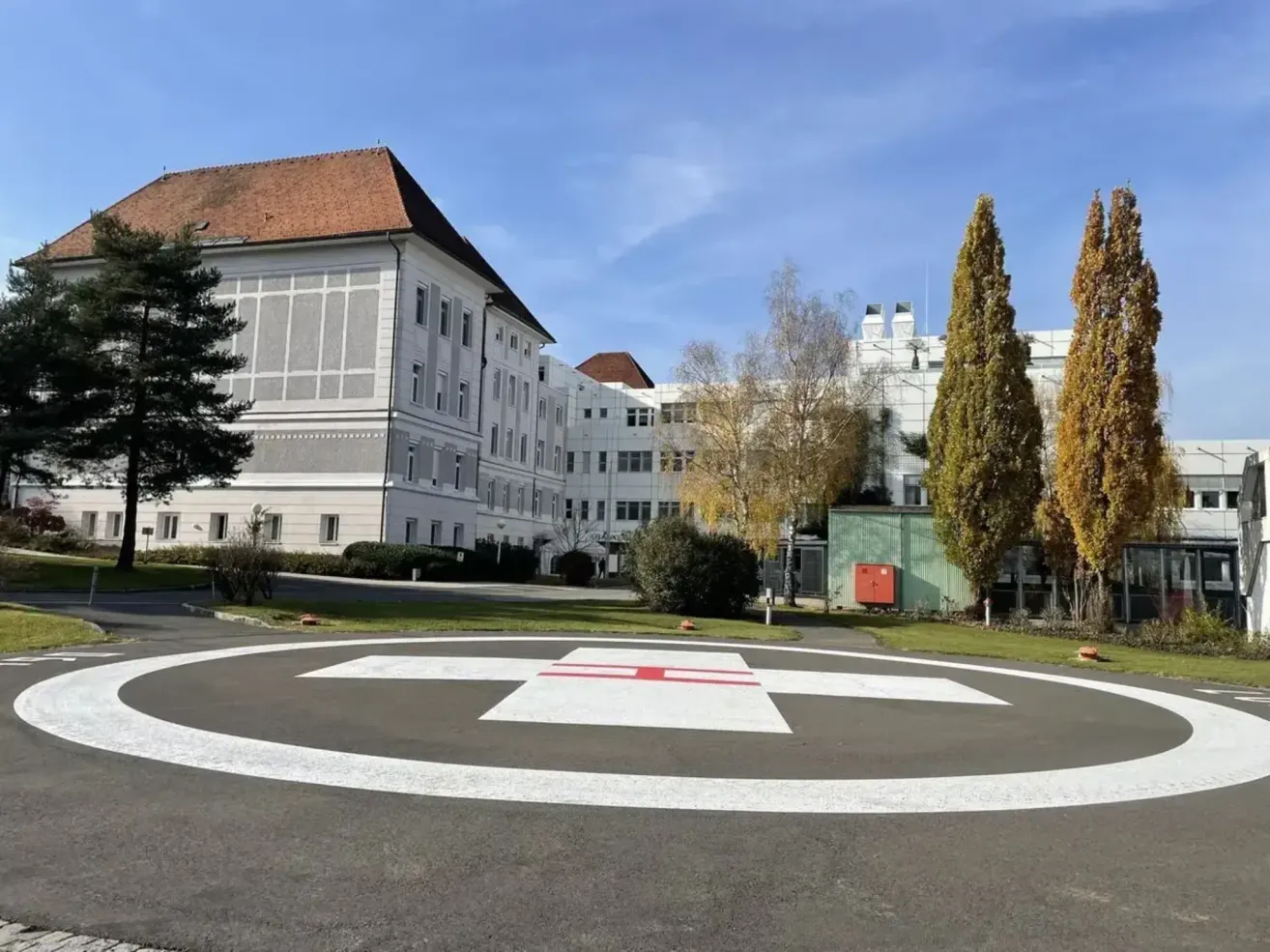 Ein großes weißes Gebäude mit rotem Dach steht hinter einem grünen Rasen. Ein weißes Kreuzsymbol markiert den Boden, und zwei Bäume sind in der Nähe.