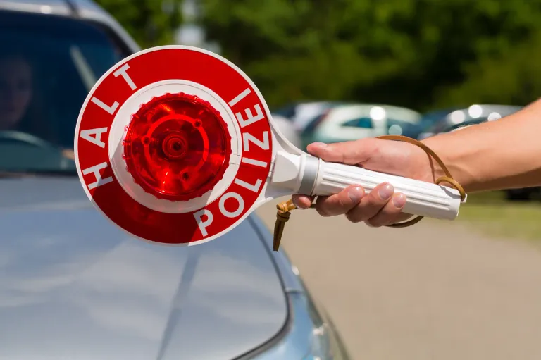 Eine Hand hält ein rotes und weißes Polizei-Warnlicht auf einem blauen Auto mit Bäumen im Hintergrund.