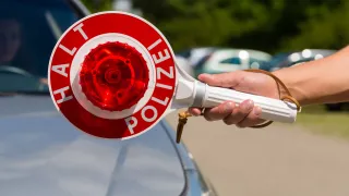 Eine Hand hält ein rotes und weißes Polizei-Warnlicht auf einem blauen Auto mit Bäumen im Hintergrund.