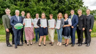 Eine Gruppe von Männern und Frauen posiert für ein Foto, während sie Urkunden halten. Sie stehen draußen an einem sonnigen Tag.