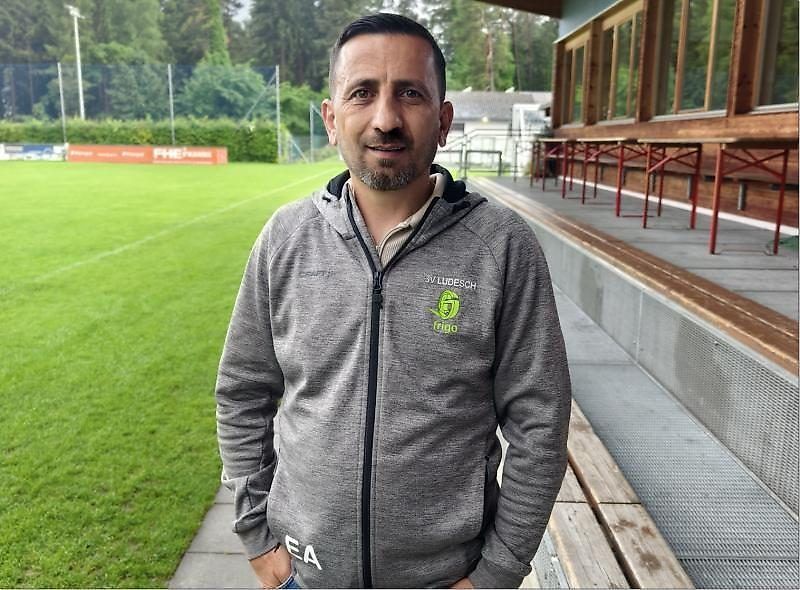 Ein Mann steht auf einem Weg vor einem Feld, trägt eine graue Kapuzenjacke mit einem Logo auf der Brust. Hinter ihm befindet sich ein grünes Feld und rote Bänke.