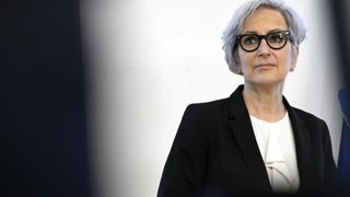 Eine Frau mit kurzen grauen Haaren, die eine Brille und einen schwarzen Blazer trägt, steht vor einer weißen Wand.