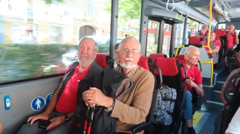 Zwei Männer sitzen in einem Bus. Einer trägt ein rotes Shirt, der andere einen braunen Pullover. Beide halten Stöcke. Der Bus ist draußen geparkt.