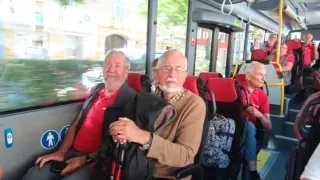 Zwei Männer sitzen in einem Bus. Einer trägt ein rotes Shirt, der andere einen braunen Pullover. Beide halten Stöcke. Der Bus ist draußen geparkt.