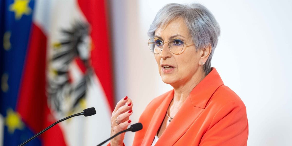 Eine ältere Frau mit grauem Haar spricht in ein Mikrofon und trägt eine Brille und einen orangefarbenen Blazer. Hinter ihr ist eine Flagge mit einem Logo zu sehen.