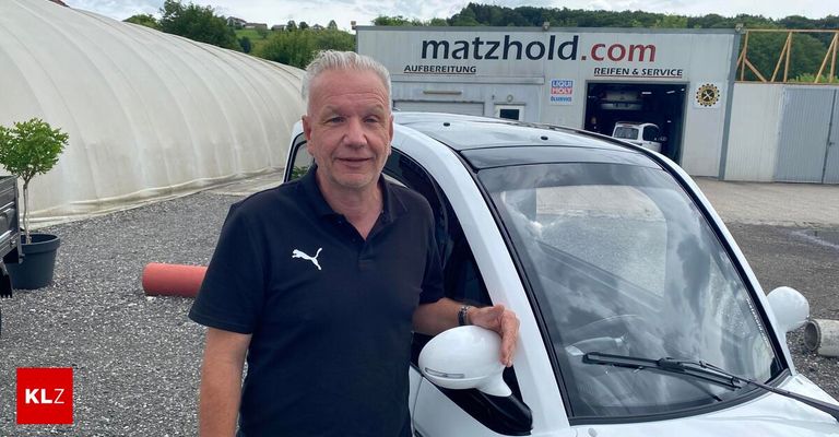 Ein Mann steht neben einem weißen Auto und hält dessen Türspiegel. Hinter ihm befindet sich ein Gebäude mit der Aufschrift 'matzhold.com'.