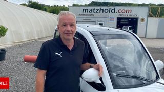 Ein Mann steht neben einem weißen Auto und hält dessen Türspiegel. Hinter ihm befindet sich ein Gebäude mit der Aufschrift 'matzhold.com'.