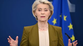 Eine Frau in einem grünen Blazer und einer Perlenkette spricht an einem Podium vor EU-Fahnen im Hintergrund.