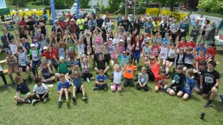 Eine Gruppe von Teilnehmern bei einem Laufevent sitzt auf dem Rasen und hebt die Hände. Sie tragen nummerierte Shirts. Im Hintergrund befindet sich ein Spielplatz.