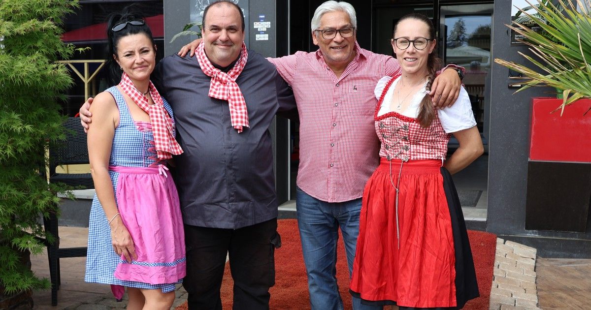 Vier Personen stehen eng zusammen in traditioneller Kleidung und lächeln für die Kamera. Der Mann links trägt ein dunkles Hemd, und der Mann rechts trägt ein rotes kariertes Hemd. Die beiden Frauen tragen rosa und blaue Kleider.