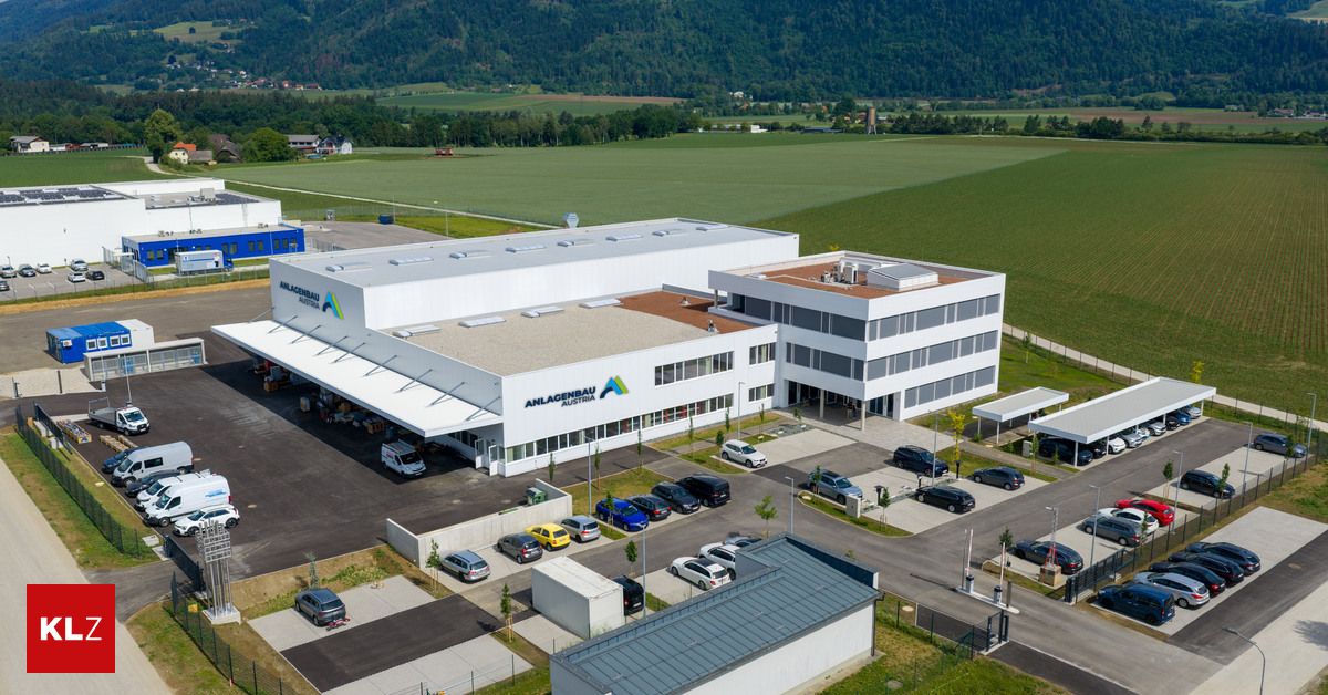 Luftaufnahme eines modernen Industriegebäudes mit mehreren geparkten Fahrzeugen davor und einem großen grünen Feld im Hintergrund.