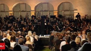 Ein Symphonieorchester spielt auf der Bühne für ein Publikum, mit Musikern, die Violinen, Celli, Trompeten und andere Instrumente spielen. Der Dirigent steht vorne in der Mitte und leitet die Aufführung.