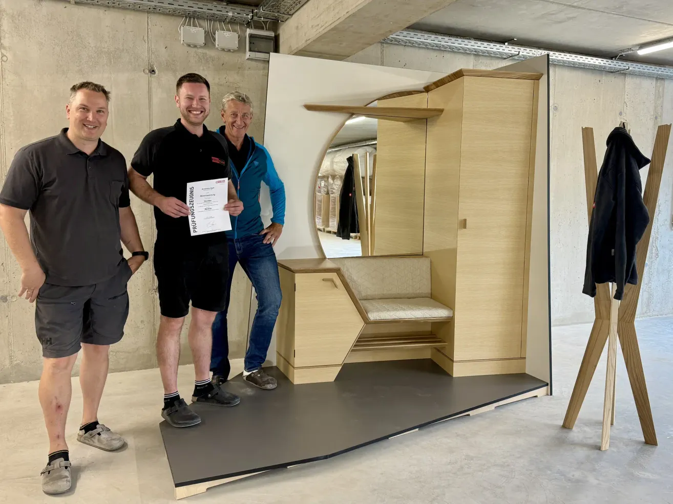 Drei Männer stehen neben einem Holzmöbelstück. Der Mann in der Mitte hält ein Zertifikat. Das Möbelstück hat eine Bank und einen Spiegel. Sie stehen auf einer Plattform mit einer grauen Matte.