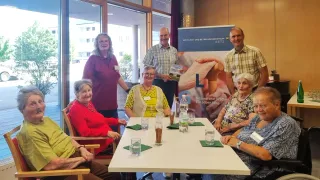 Eine Gruppe älterer Menschen, darunter einige Frauen, sitzen um einen Tisch mit Wassergläsern, Servietten und einem Banner hinter ihnen. Es stehen auch zwei Männer hinter ihnen.