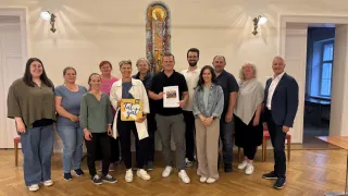 Eine Gruppe von Menschen steht in einem Raum mit Holzboden. Sie posieren für ein Foto. Eine Frau hält ein Schild mit der Aufschrift Tut gut! Ein Mann hält ein Papier. Dahinter befindet sich eine Wand mit einem Buntglasfenster.