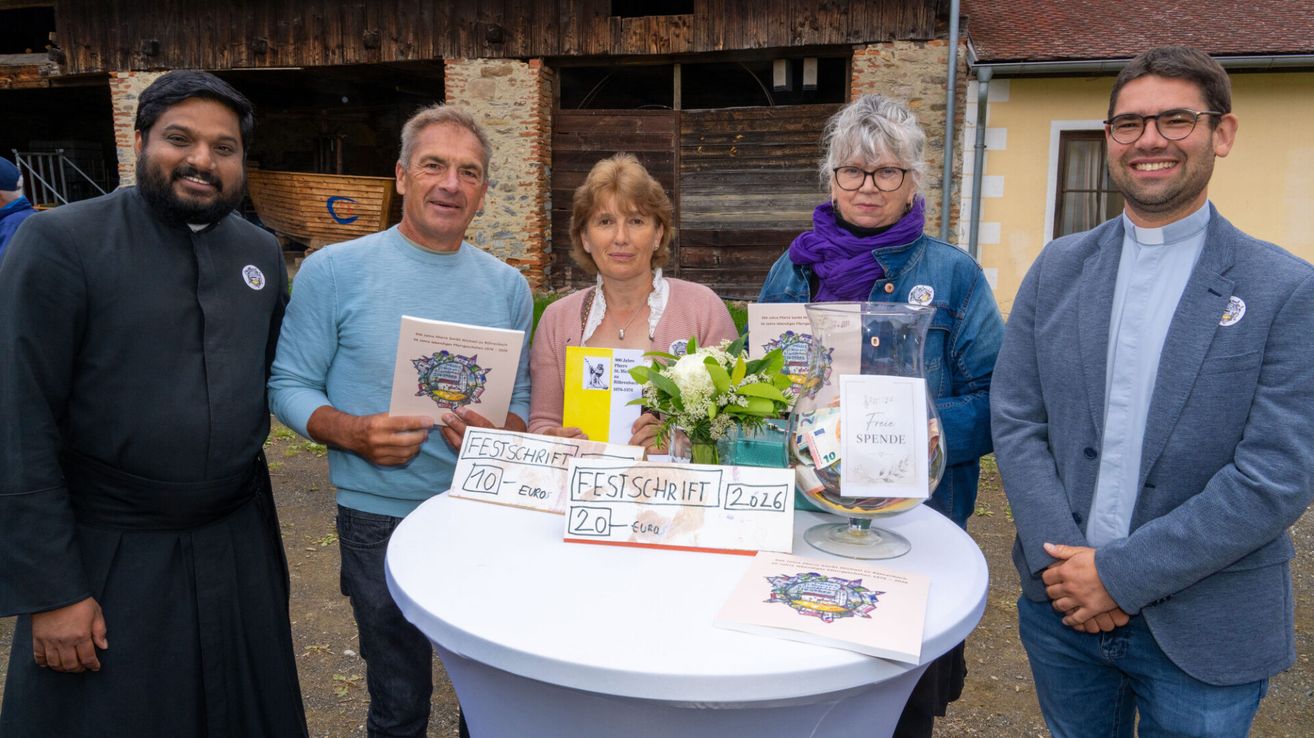 Drei Personen stehen hinter einem Tisch mit Blumen und Broschüren und halten Broschüren mit Nummern in der Hand. Sie scheinen zu lächeln und für ein Foto zu posieren.