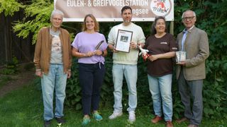 Vier Personen stehen in einem Grasbereich mit einem Banner hinter ihnen. Ein Mann hält ein Zertifikat und eine Feder. Die Frau neben ihm hält eine Feder.