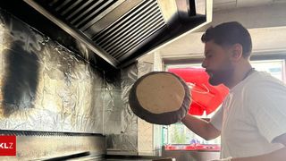 Ein Mann kocht eine Tortilla auf einem Grill in einer Restaurantküche, wobei ein roter Schirm vor dem Fenster zu sehen ist.