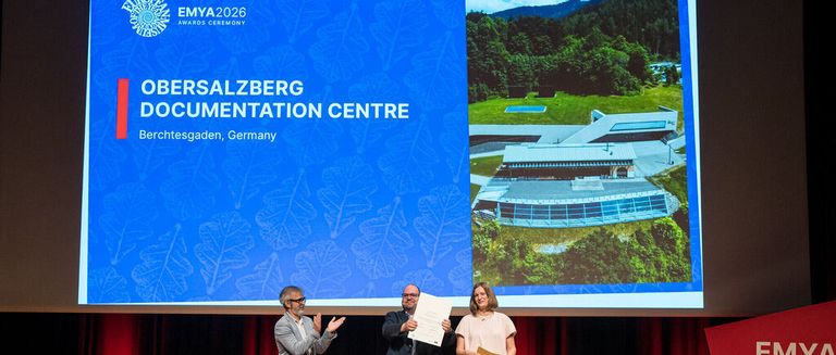 Three individuals stand on stage with the Obersalzberg Documentation Centre in the background, one holding a paper and two holding certificates.