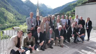 Eine Gruppe von Menschen in Geschäftskleidung und Umhängebändern posiert für ein Foto vor Bergen und einer Kirche.
