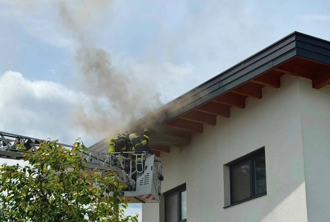 Feuerwehrleute auf einer Leiter an einem Gebäude, mit Rauch, der vom Dach aufsteigt. Das Gebäude hat eine weiße Wand, ein braunes Dach und zwei Fenster.