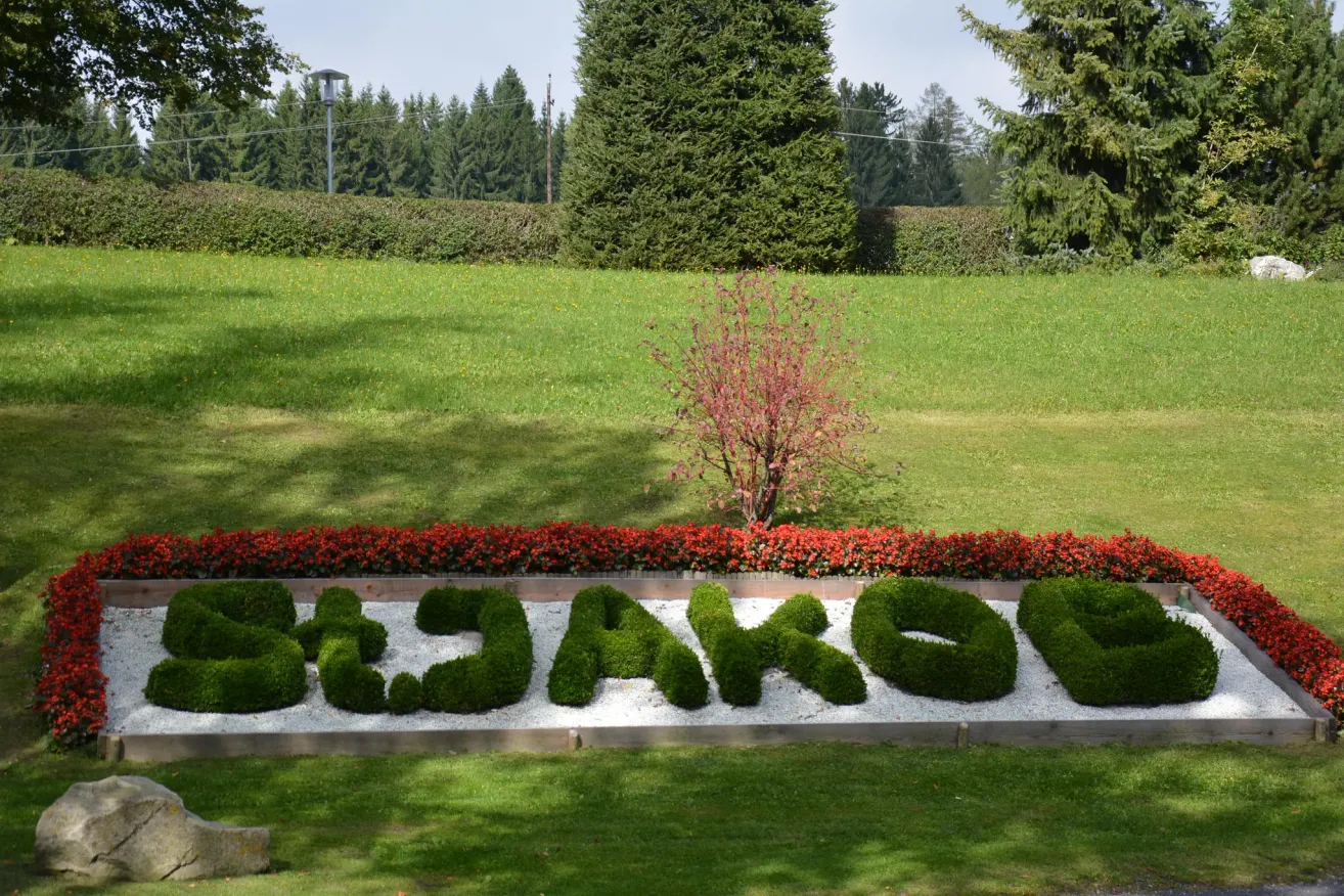 Das Wort 'BJAKON' ist kunstvoll aus grünen Sträuchern gestaltet. Es ist von einem Rand aus roten Blumen umgeben. Ein grasbewachsenes Feld mit Bäumen und Büschen befindet sich im Hintergrund.