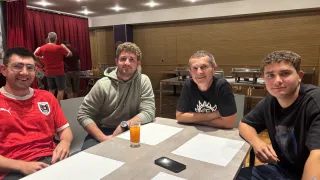 Vier Männer sitzen an einem Tisch in einem Raum. Ein Mann trägt eine graue Kapuzenjacke, und ein anderer trägt ein schwarzes T-Shirt mit einem weißen Design. Ein Glas Orangensaft steht auf dem Tisch.