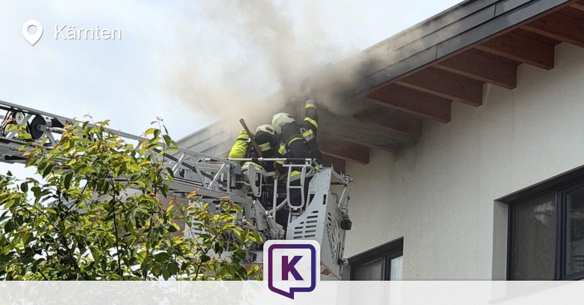 Feuerwehrleute stehen auf einer Leiter und arbeiten daran, ein Feuer auf dem Dach eines Gebäudes zu löschen. Rauch steigt von der Szene auf.