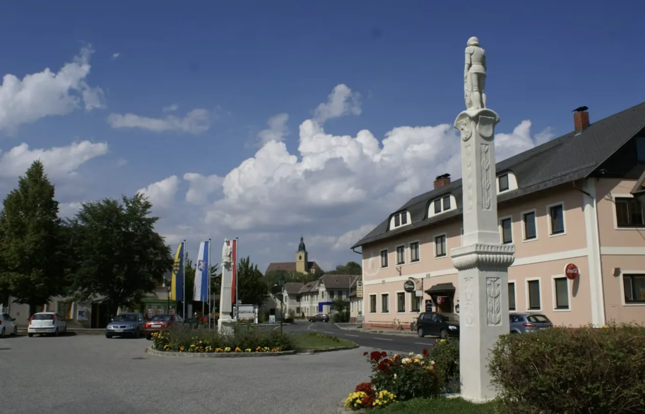 Ein Stadtplatz mit einer hohen Statue in der Mitte. Mehrere Flaggen und Denkmäler sind vorhanden. Der Himmel ist blau mit Wolken. Autos und Gebäude säumen die Straße.