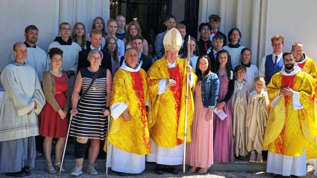 Eine Gruppe von Menschen steht in einer Reihe vor einer Kirche. Ein Bischof und ein Priester tragen gelbe und rote Roben. Sie stehen vor der Gruppe. Die Menschen tragen formelle Kleidung.