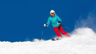 Ein Skifahrer in einer blauen Jacke und roten Hosen rutscht einen verschneiten Berg hinunter. Der Skifahrer trägt einen weißen Helm, eine Sonnenbrille und Handschuhe.